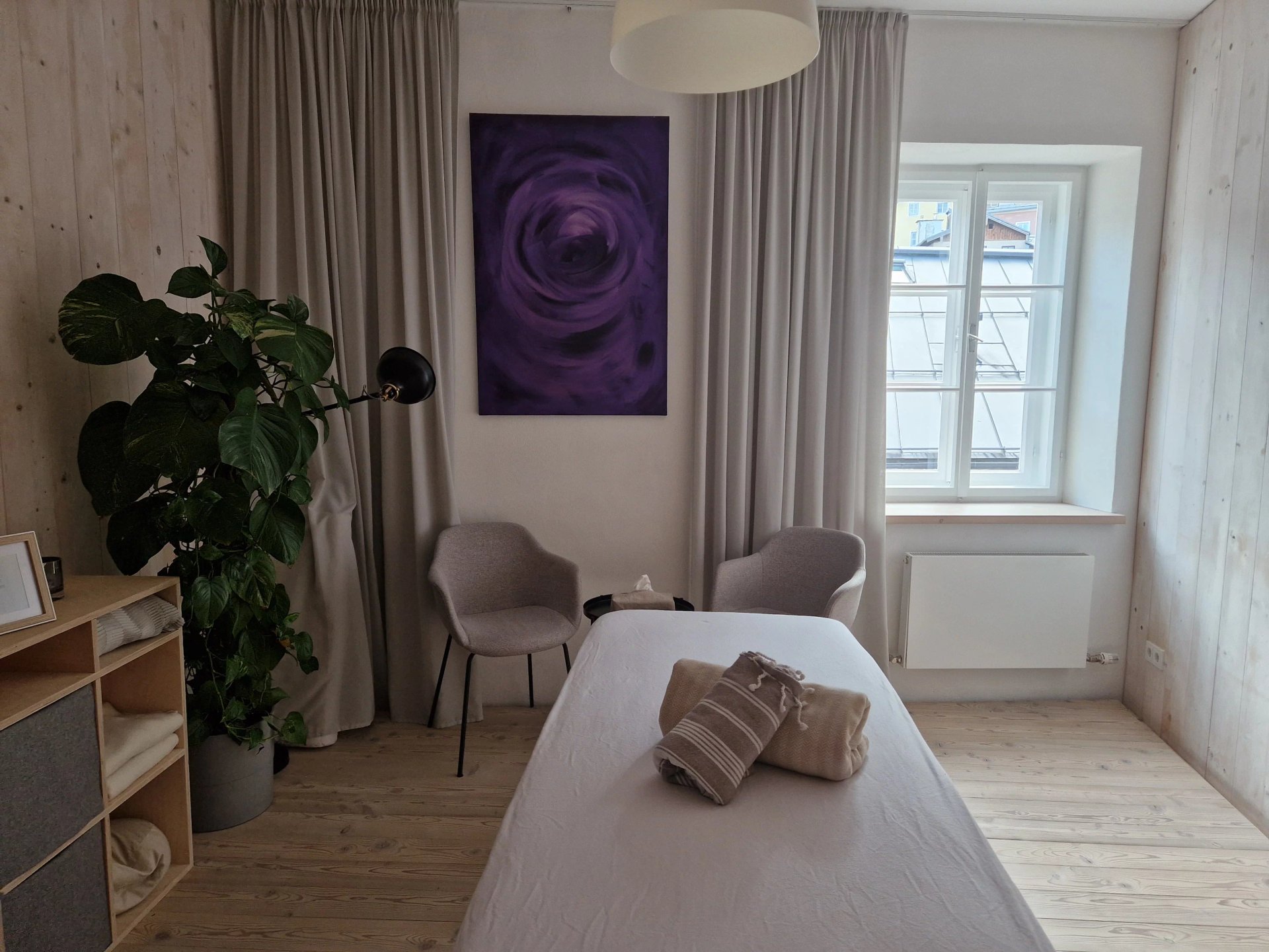 Eine Massageliege in einem gemütlichen, hellen Raum mit Holzboden und umrahmt von einer Pflanze und einer Sitzgruppe.
