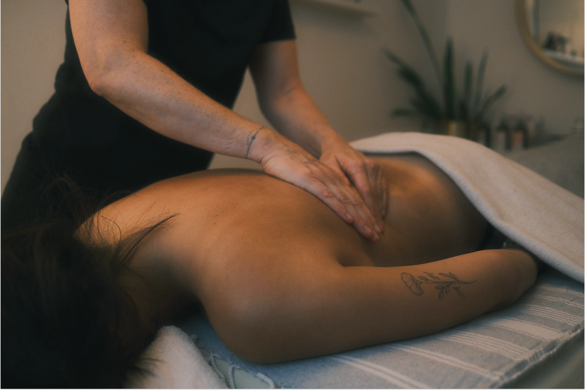 Eine Person liegt mit nacktem Rücken auf einer Massageliege, während Sabine ihr mit beiden Händen den Rücken massiert. Die empfangende Person ist teilweise mit einer Decke bedeckt. Der Raum wirkt warm und beruhigend